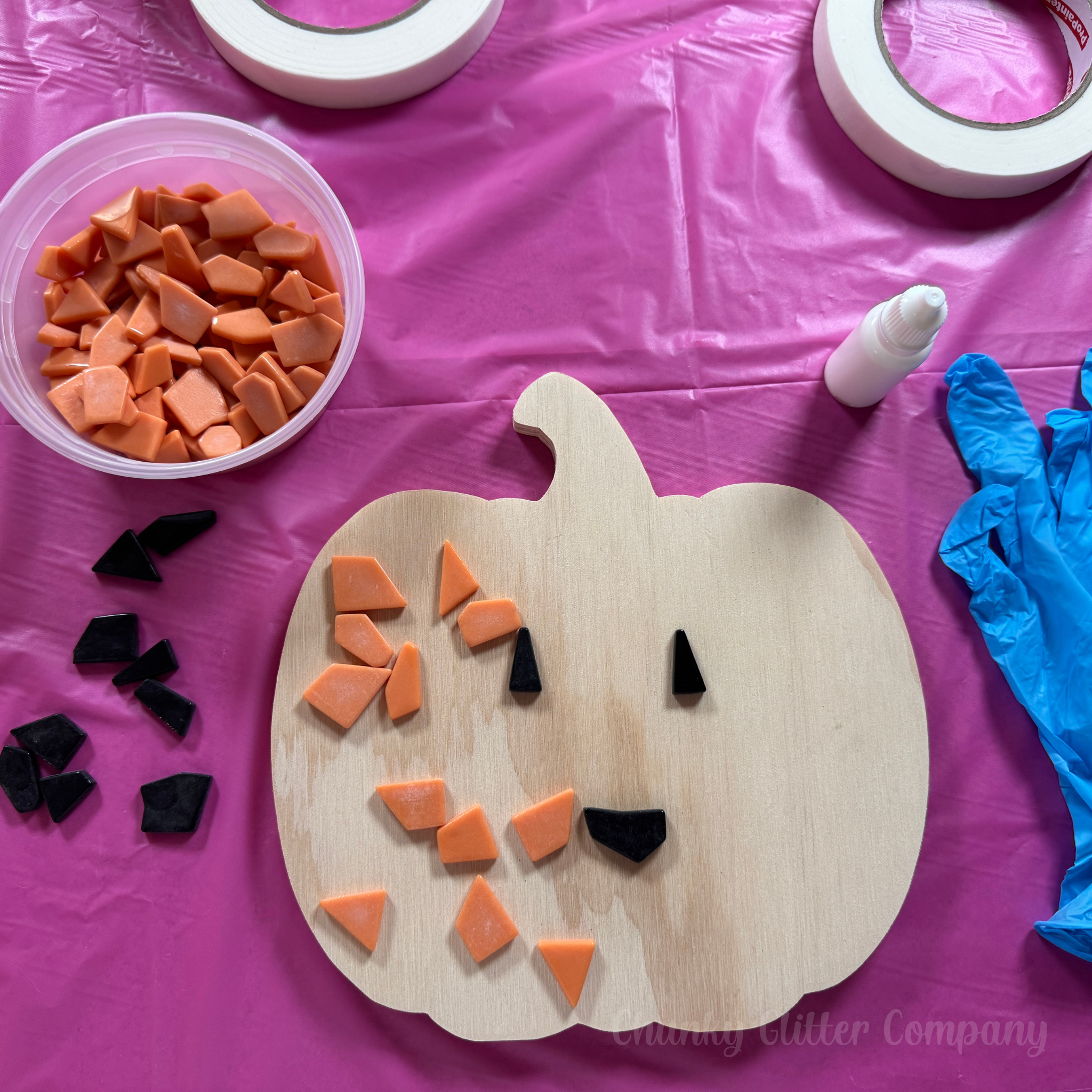 Wooden pumpkin decoration with orange and black pieces on a pink table cloth, with tape and gloves nearby.