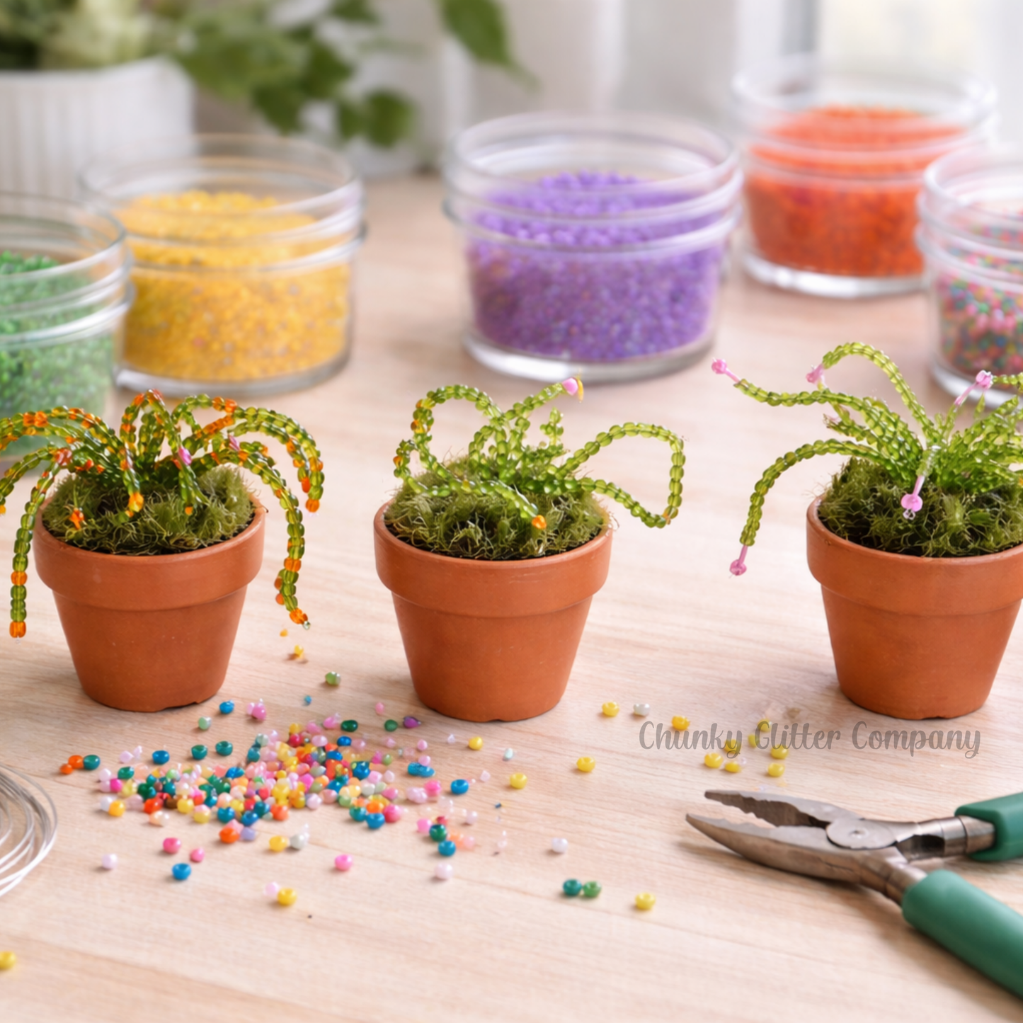 Three small beaded plants with decorative beads on a wooden surface, surrounded by bead containers and pliers.