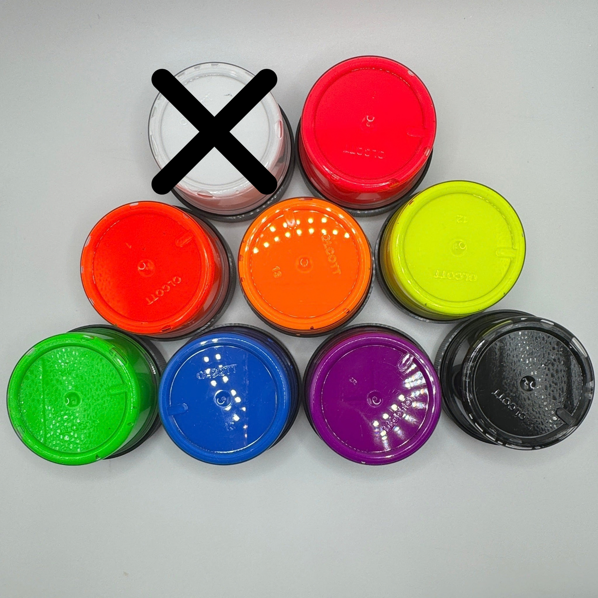 Nine small round plastic tubs with colorful lids arranged in a pyramid on a light gray background. The top-left tub is crossed out with a large black X, indicating it is excluded from the color selection.