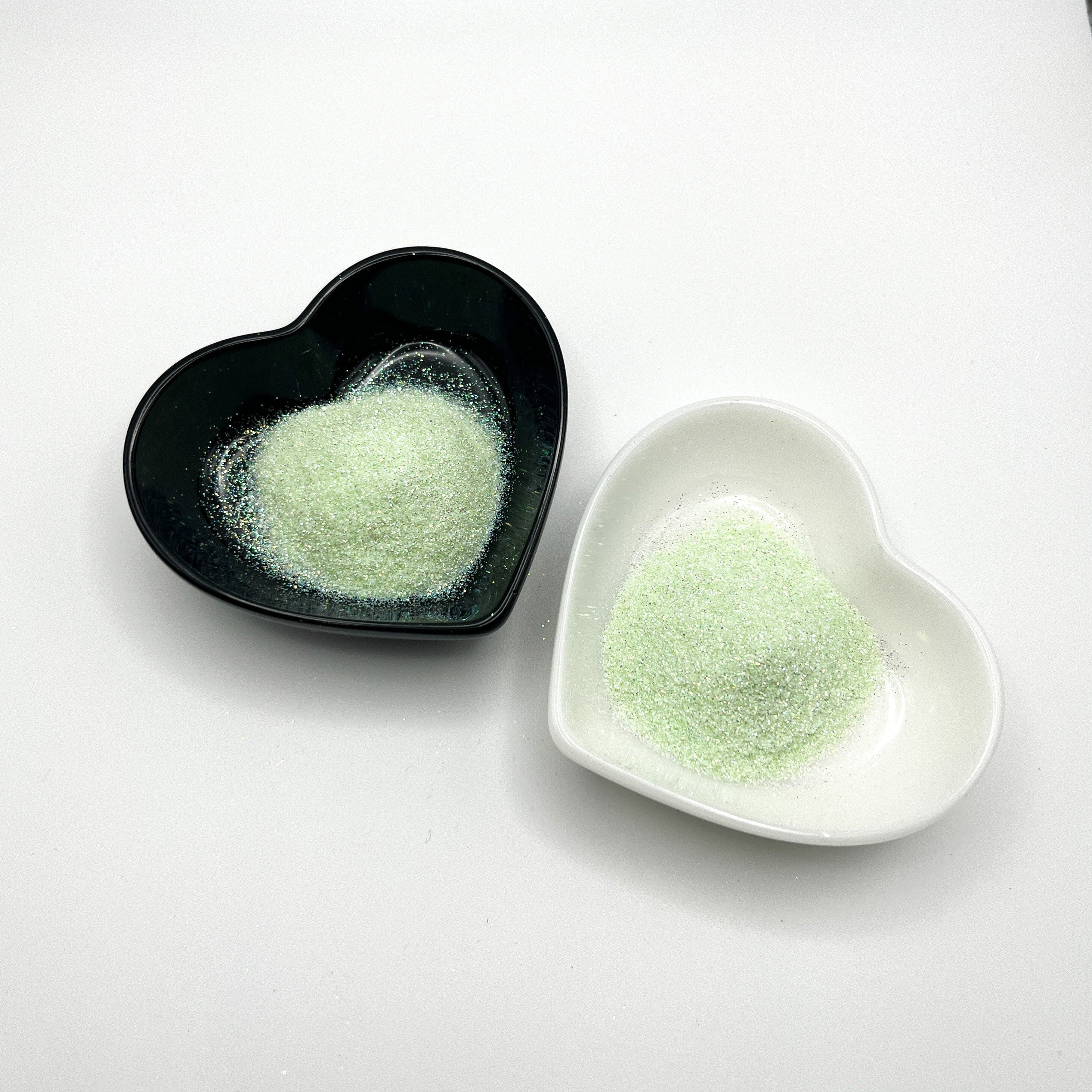 Two heart-shaped dishes, one black and one white, both containing pale green extra fine glitter.