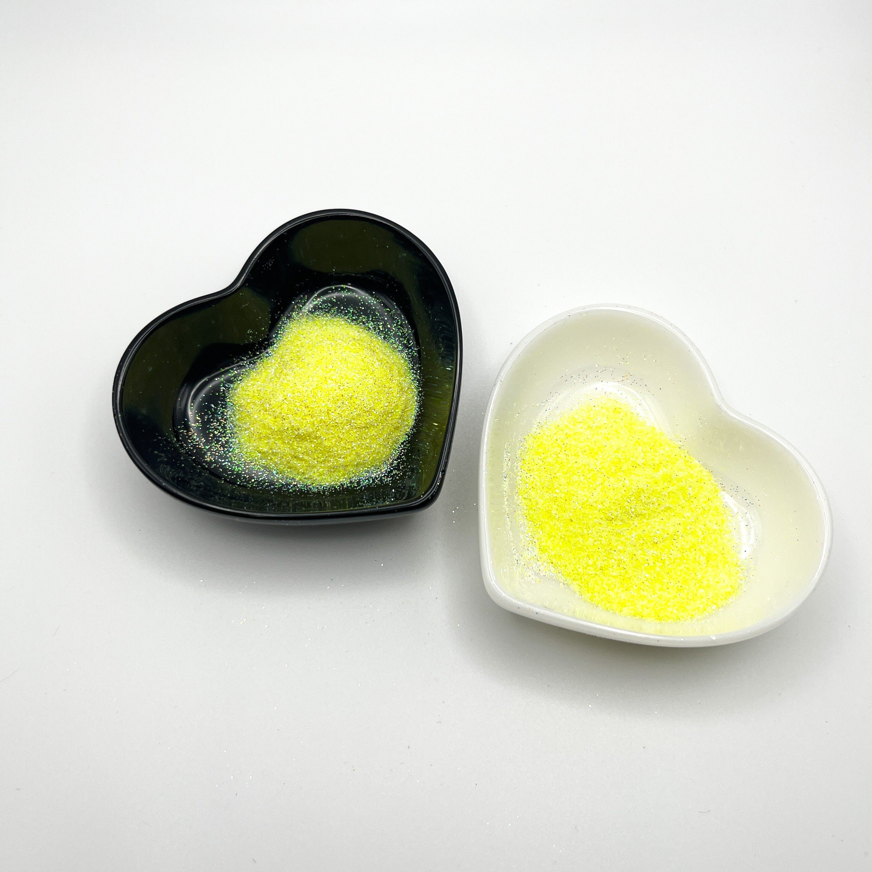 Two heart-shaped dishes, one black and one white, both containing pale yellow extra fine glitter.