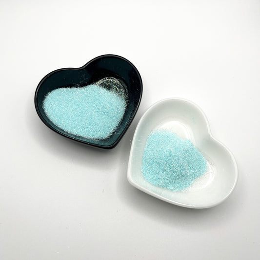 Two heart-shaped dishes, one black and one white, both containing light blue extra fine glitter.