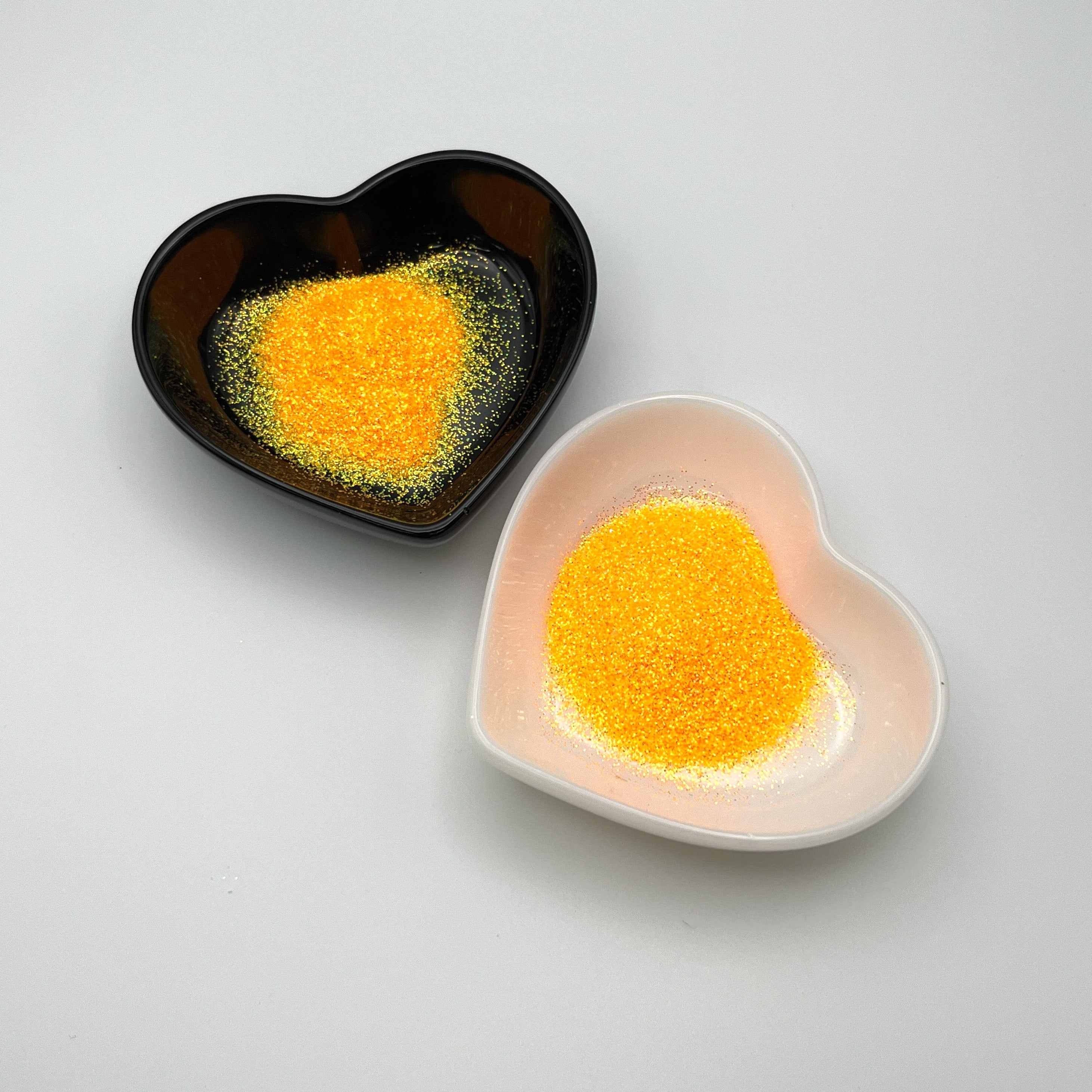 Two heart-shaped dishes, one black and one white, both containing orange extra fine glitter.