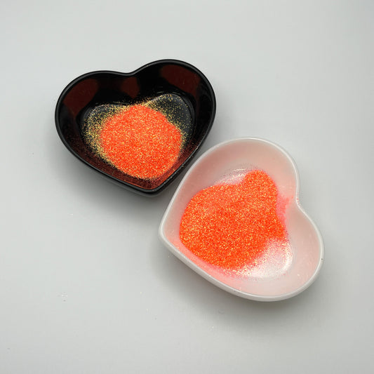 Two heart-shaped dishes, one black and one white, both containing coral extra fine glitter.