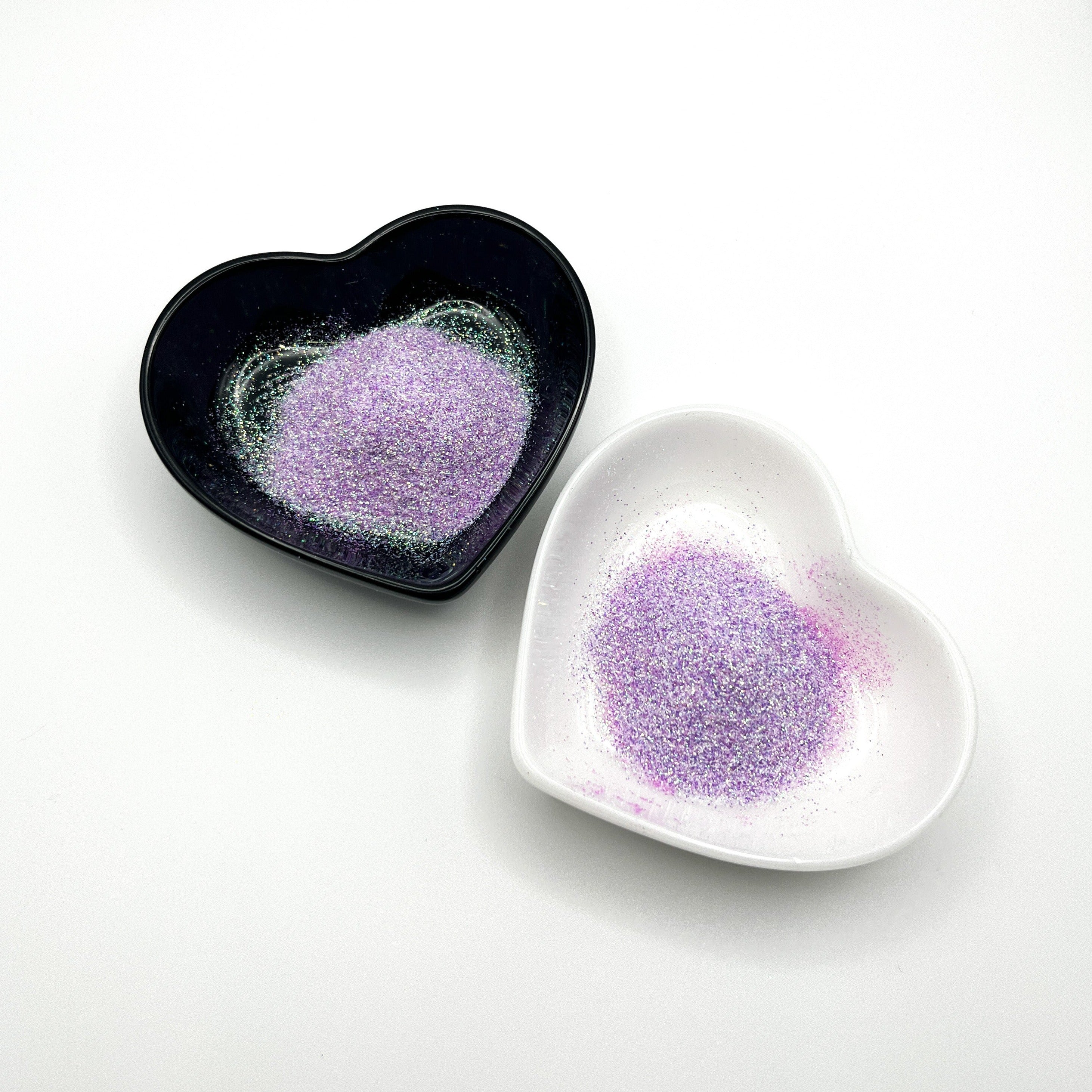 Two heart-shaped dishes, one black and one white, both containing light purple extra fine glitter.
