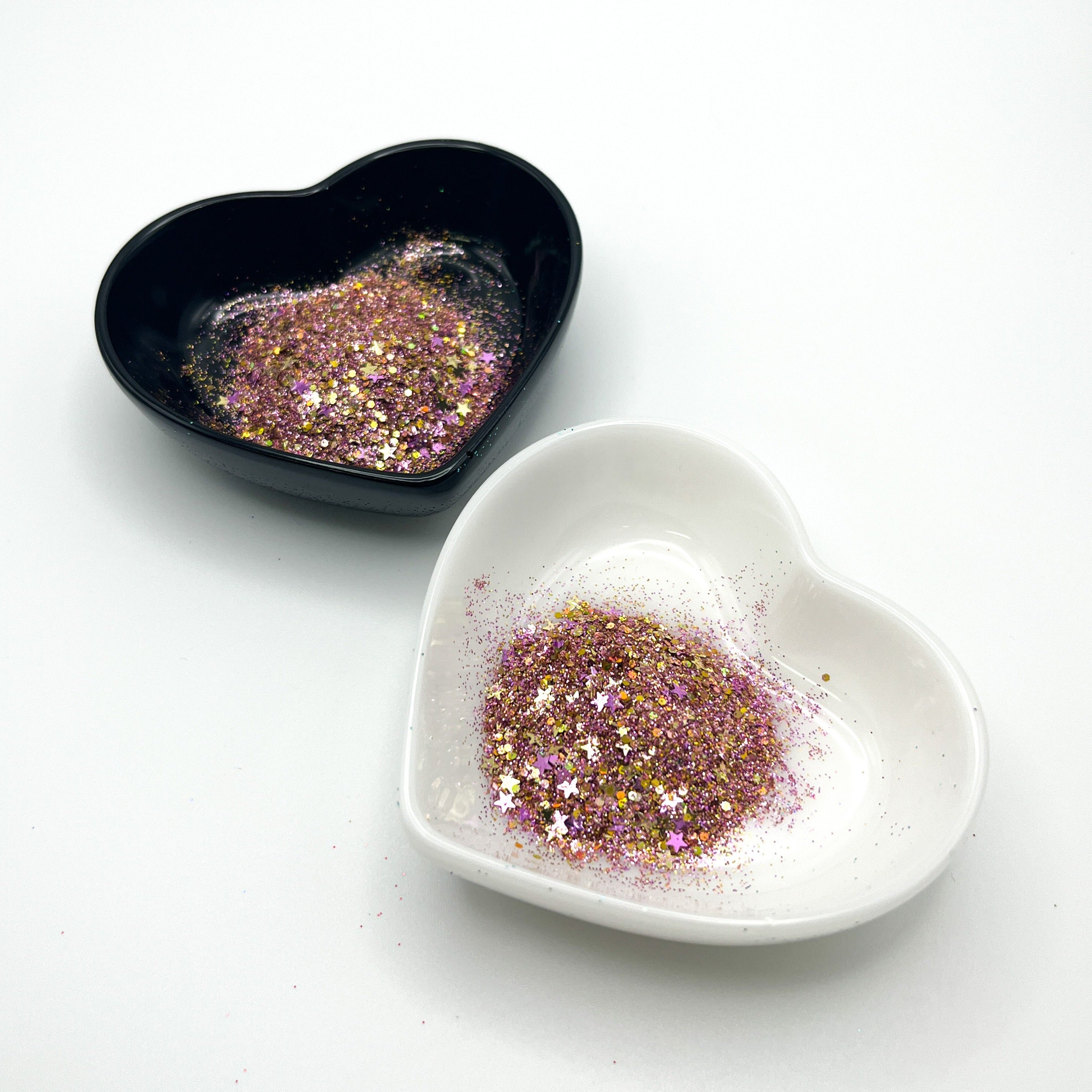 Two heart shaped containers, one black, one white, containing a Gold/Purple chunky glitter mix with hexagons and stars.