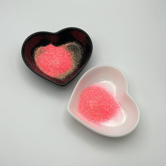 Two heart-shaped dishes, one black and one white, both containing pink extra fine glitter.