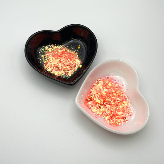 Two heart-shaped dishes, one black and one white, containing pink and yellow chunky glitter mix.