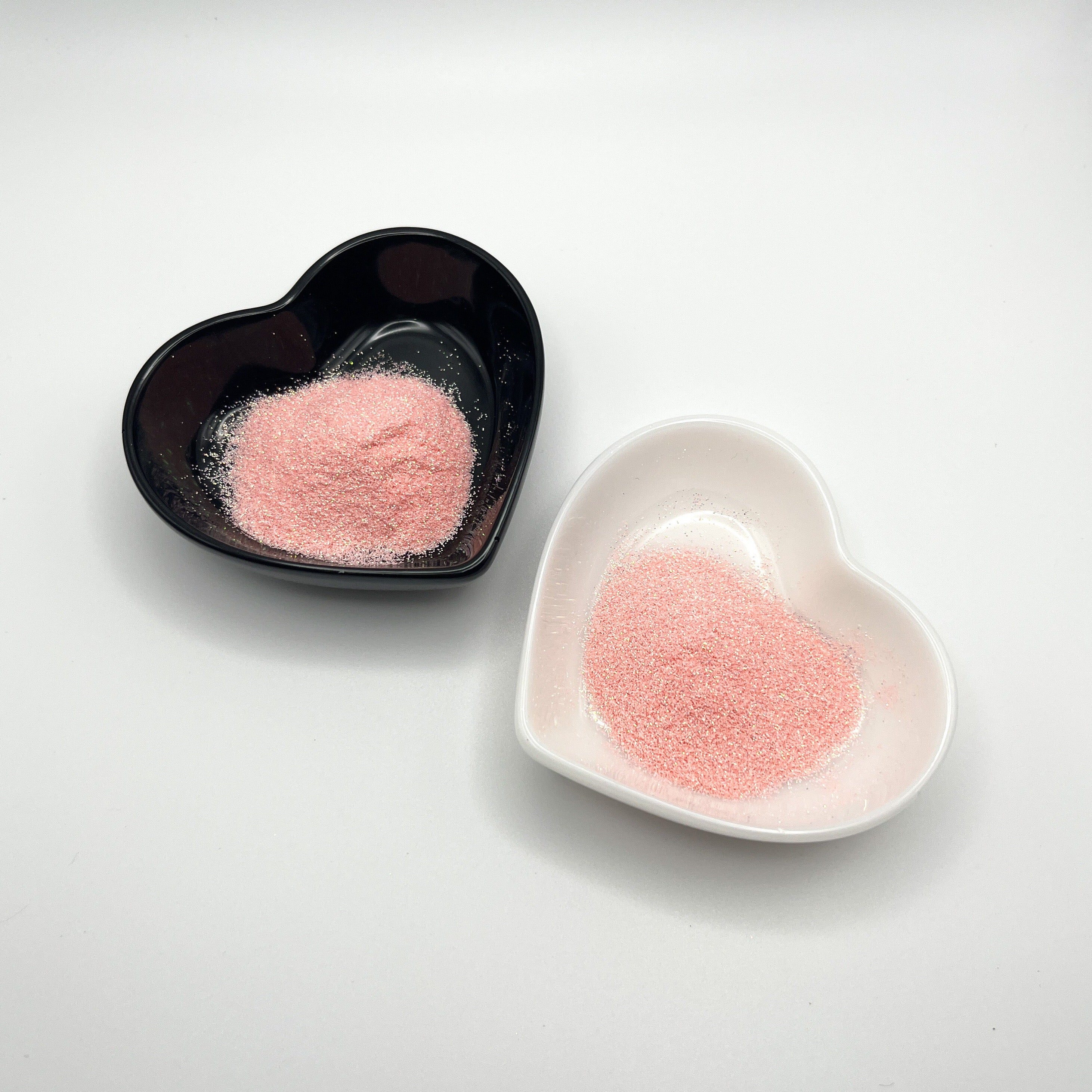 Two heart-shaped dishes, one black and one white, both containing light pink extra fine glitter.