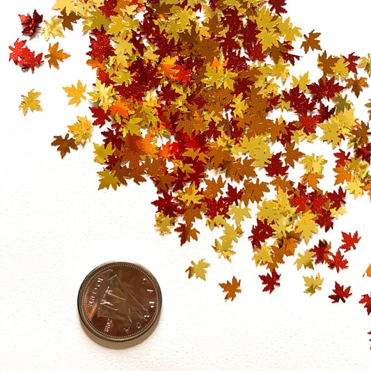 A collection of orange, red, and gold colored leaf-shaped glitter pieces spread out on a surface with a dime for size reference. 