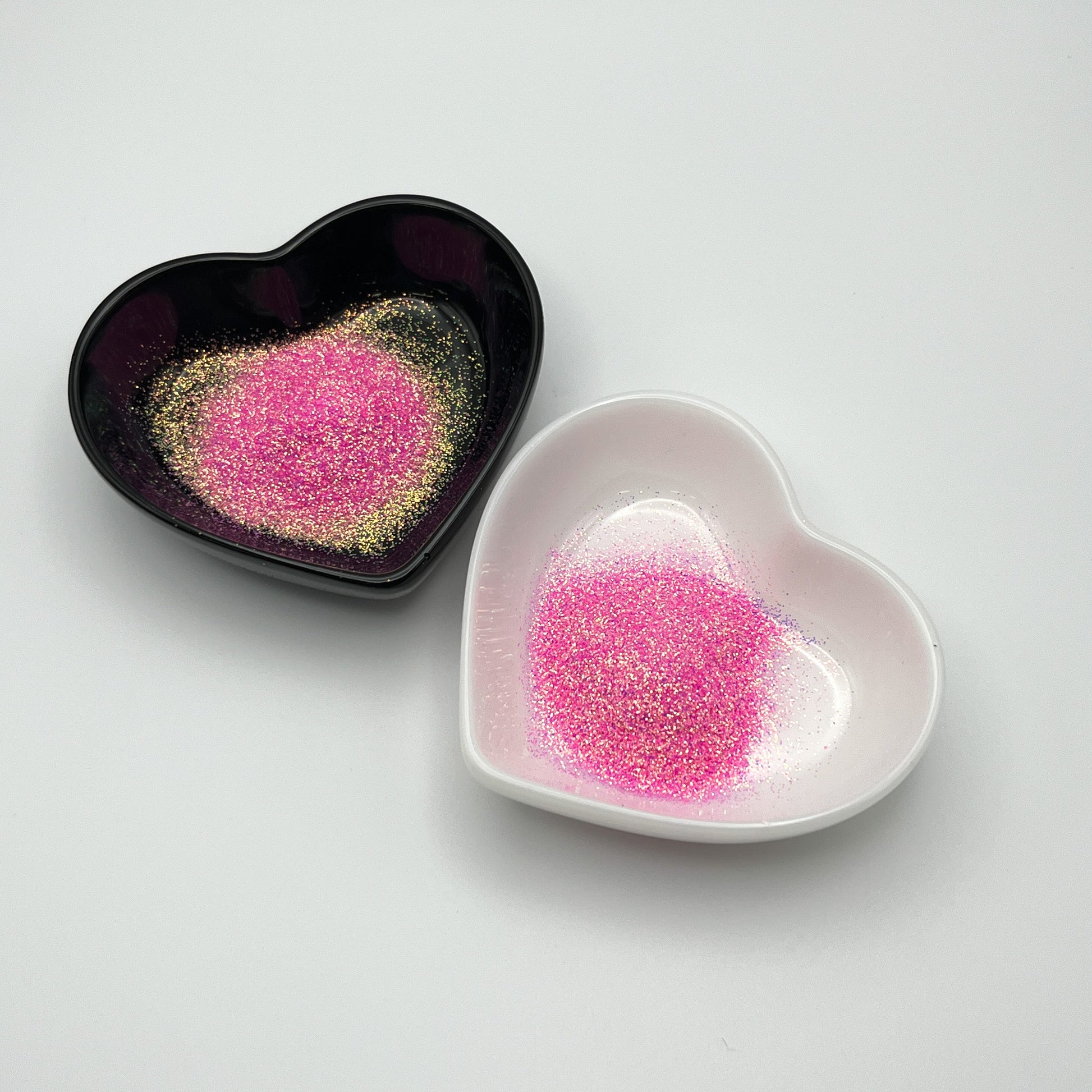 Two heart-shaped dishes, one black and one white, both containing light pink extra fine glitter.